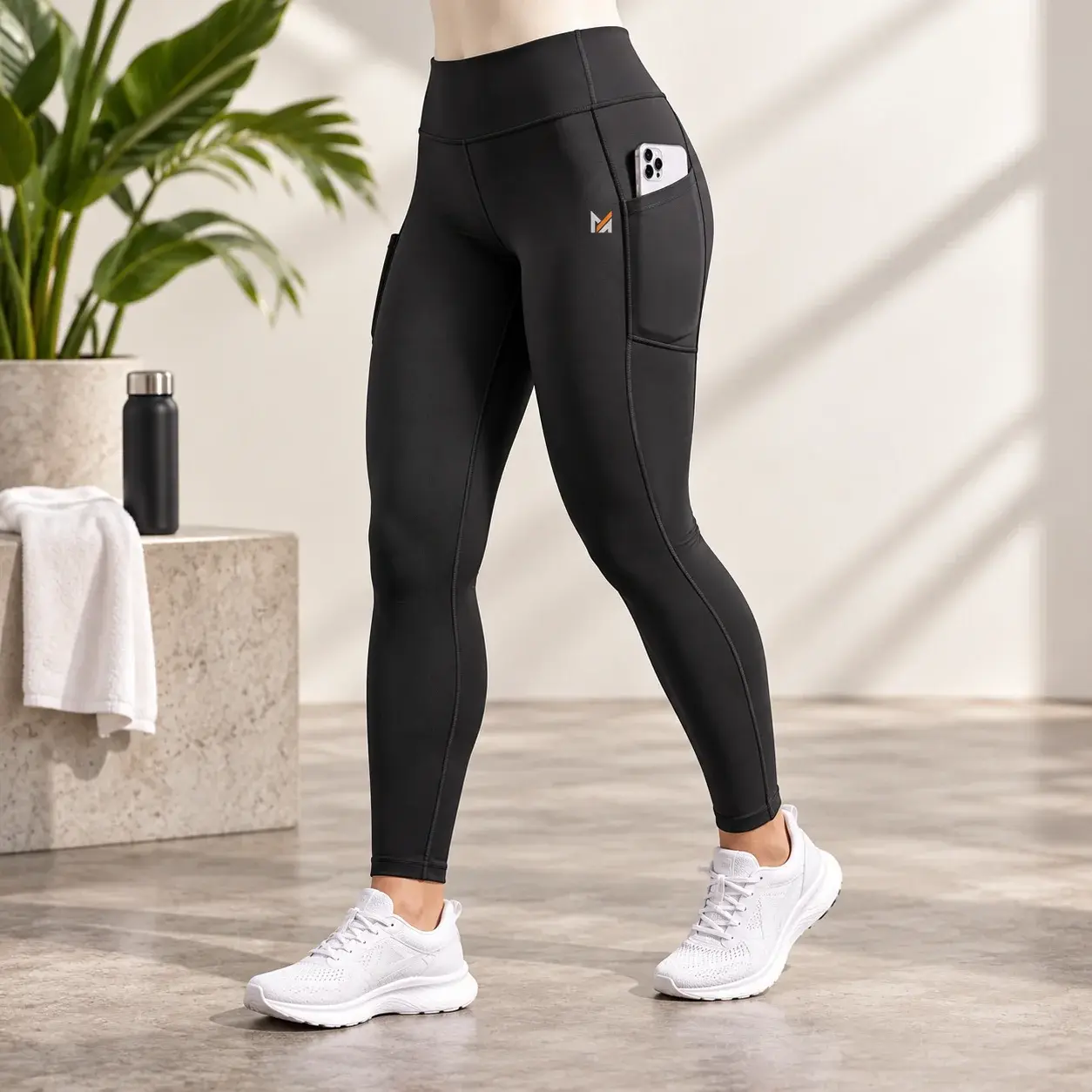 Black high-waisted performance leggings with large side pockets, smartphone storage, and a small Mega Apparel logo, displayed in a bright modern fitness studio.